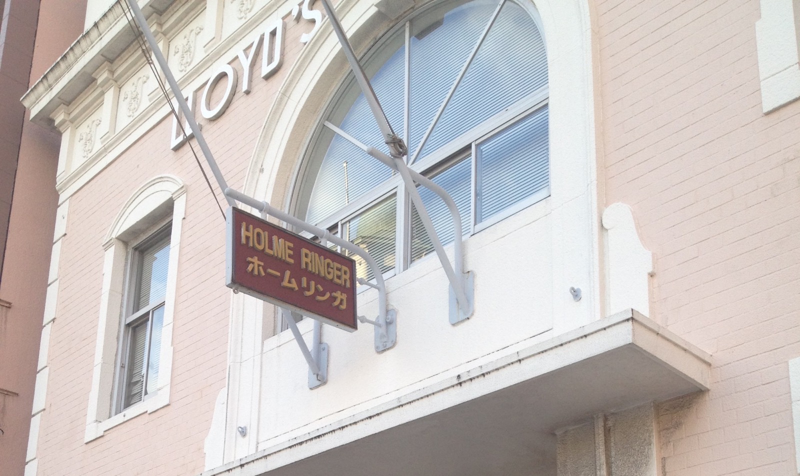 A hanging sign reading HOLME RINGER (and the same name in katakana) in front of the pink brick building in Mojiko whose facade carries the word LLOYD'S