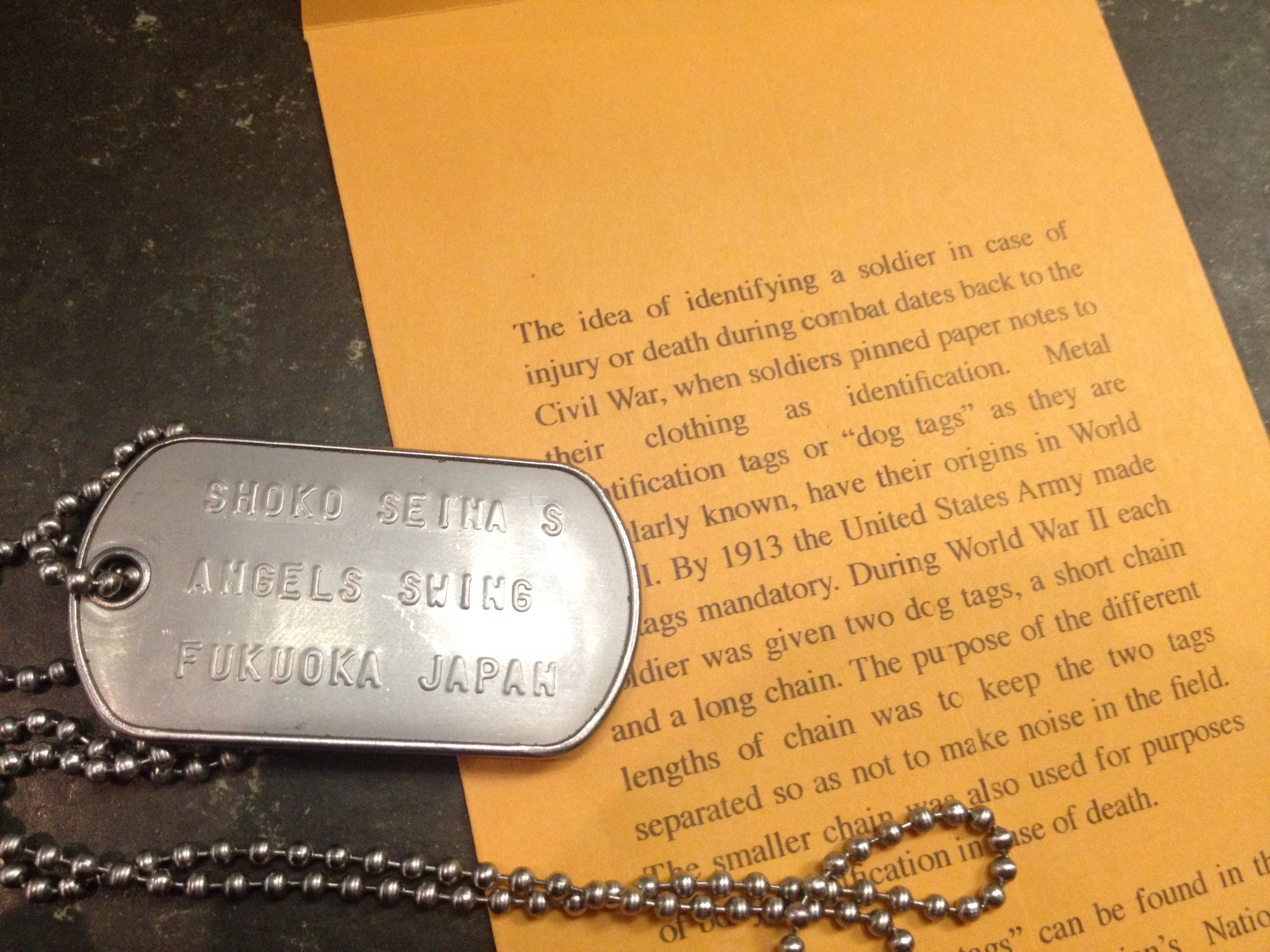 A dog tag stamped SHOKO SEINA S, ANGELS SWING, FUKUOKA JAPAN, beside a leaflet on the history of dog tags